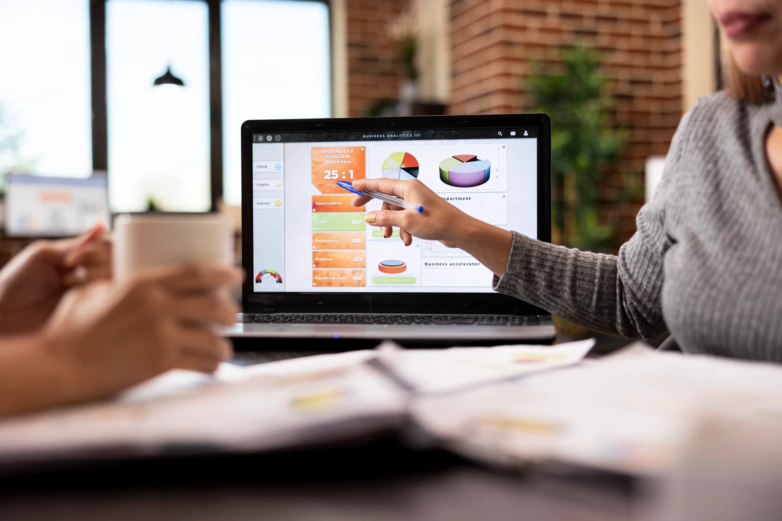 Person pointing at business analytics charts on a laptop screen in a collaborative office setting with papers and coffee.